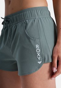 Shorts verts en tissu léger, dotés d'une ceinture élastique, de poches latérales et d'un logo blanc "ROXY" sur la jambe gauche.