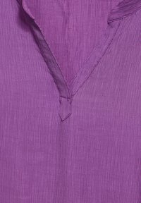 Tessuto viola texturizzato con scollatura a V e piccolo dettaglio con fiocco. Il motivo presenta strisce verticali, aggiungendo profondità al capo.