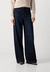 Jeans de mezclilla oscura de pierna ancha y cintura alta, con bolsillos frontales y costuras en contraste. Combinados con un suéter de punto beige y zapatos marrones.