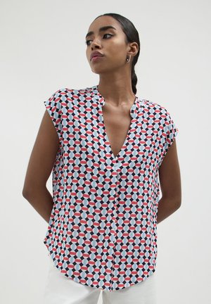 Donna che indossa un top senza maniche con un motivo geometrico a cubo blu, bianco, rosso e nero, abbinato a pantaloni bianchi, in piedi con le mani dietro la schiena.
