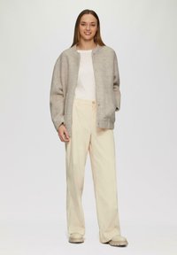 Lichtgrijze oversized cardigan met knoopsluiting, gecombineerd met crèmekleurige broek met wijde pijpen en beige schoenen. Soepele textuur en ontspannen pasvorm.