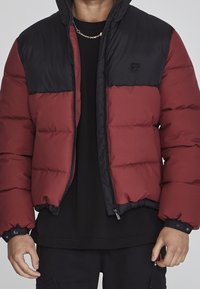 Rød og sort dunjakke med lynlås foran, let quiltet tekstur og logo på brystet. Har hætte og elastiske manchetten.