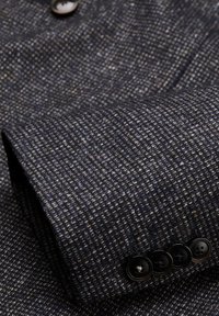 Gewebter Blazer aus dunkelblauem Stoff mit einem strukturierten Muster. Verfügt über vier schwarze Knöpfe am Ärmel mit Fadenakzenten.