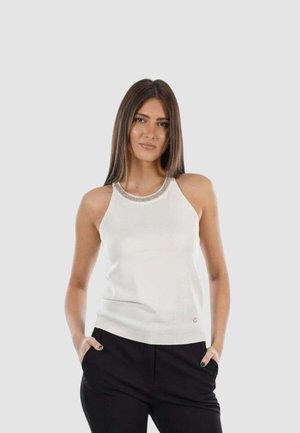 Donna con capelli castani lisci che indossa un top bianco senza maniche con dettaglio argento sul collo e pantaloni neri, mani in tasca, sfondo neutro.