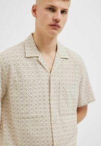 Korte mouwen shirt in lichtbeige met een bloemmotief. Heeft een zak en knoopsluiting. Gemaakt van zachte, lichtgewicht stof.