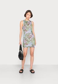 Vestido sem mangas com padrão de flores em verde e roxo sobre um fundo claro, acompanhado de sandálias pretas de slip-on e uma pequena bolsa.