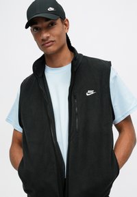 Vesta neagră din fleece cu un buzunar cu fermoar, purtată peste un tricou bleu deschis. O sapcă neagră Nike completează ținuta. Logo-ul alb Nike pe ambele articole.