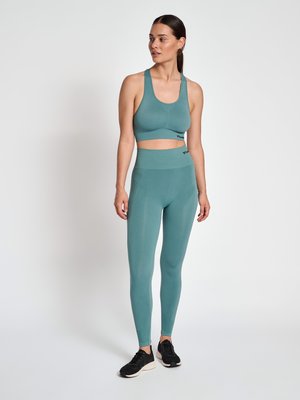 Vrouw staat, draagt een turquoise sportbeha en hoog getailleerde legging met zwarte sportschoenen, kijkt iets naar links.