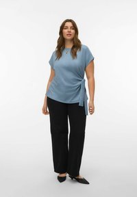 Haut bleu à manches courtes avec un détail noué sur le côté, associé à un pantalon noir large et des chaussures pointues noires, sur un fond clair uni.