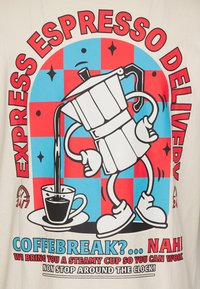 Hvid T-shirt med et tegneseriekaffemaskine, der hælder kaffe i en kop, med fed rød og blå tekst i retro stil.