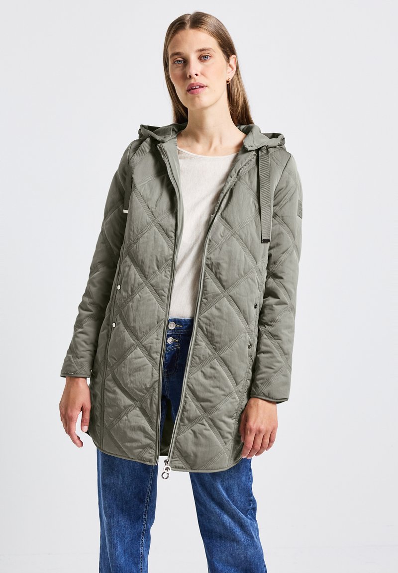 Veste matelassée verte avec capuche, boutons-pression et fermeture éclair ; présente un motif diagonal et des manches longues. Portée sur un haut léger.
