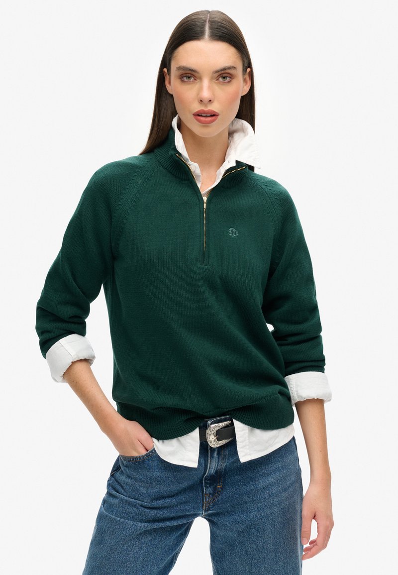 Groene gebreide trui met een kwart rits, geribbelde manchetten en een klein logo op de borst, gelaagd over een witte blouse met kraag. Jeans met een riem.