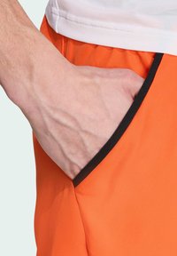 Pantalones cortos deportivos naranjas con un bolsillo lateral con detalle en negro, hechos de un tejido texturizado y ligero.