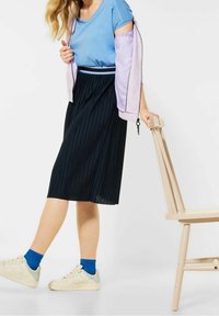 Kláčená černá midi sukně, spárovaná s modrým topem a světle fialovou bundou. Modelka má na sobě bílé tenisky a modré ponožky, drží židli.