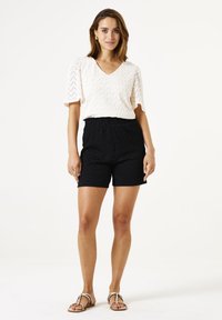 Haut V en col blanc texturé à manches courtes associé à un short noir à taille élastique avec un motif. Sandales plates beiges.