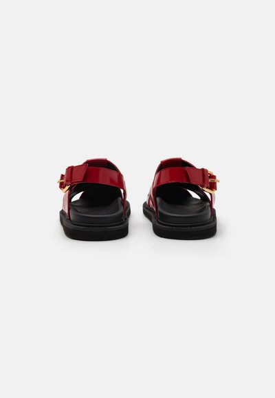 Punaiset patenttinateksiset sandaalit, joissa on tuplat säädettävät soljet, mustat kumipohjat ja avoin takaosa. Litteä, teksturoitu pohja mukavuutta varten.
