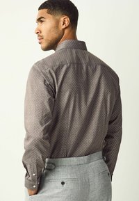 Men's shirt in dark brown fabric with light polka dots. Features a classic collar, long sleeves, and button cuffs. Paired with grey trousers.