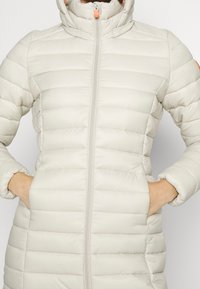 Lys beige polstret jakke med høj hals, lynlåslukning og to forlomma. Har vandret syninger og en orange lynlåsgreb.