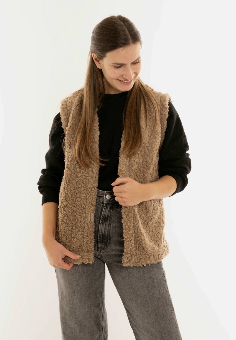 Faux fur mouwloos vest in lichtbruin, met een gestructureerd oppervlak. Gemaakt over een zwart langarmshirt en gecombineerd met hoge taille, donkere jeans.