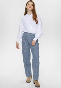 Chemise blanche à manches longues et poignets retroussés, associée à un pantalon large à rayures bleues et blanches et des bottines beiges.