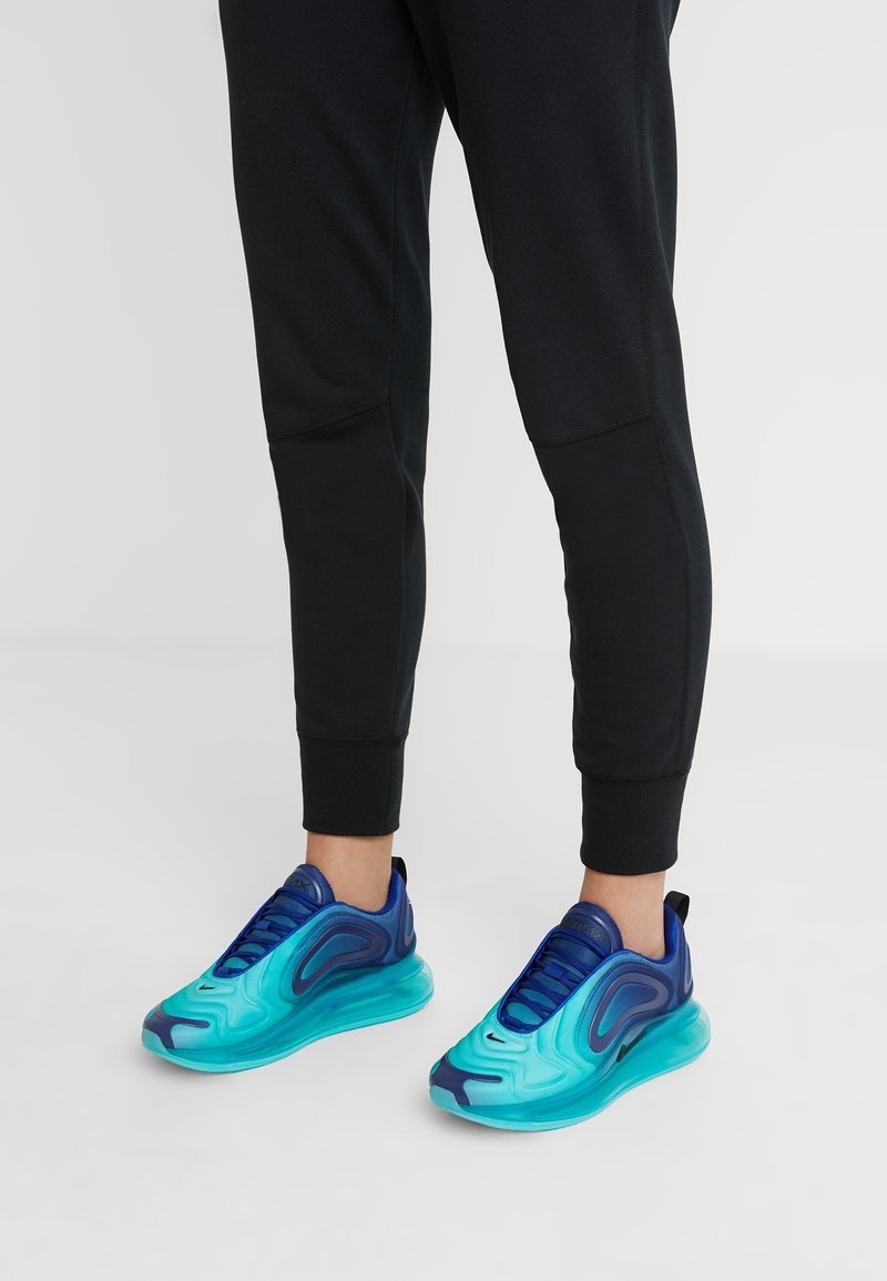 Personne portant un jogging noir et des baskets bleu vif et turquoise, debout sur un sol gris clair avec un arrière-plan uni.