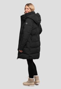 Zwarte pufferjas met een capuchon, voorzien van een doorgestikte ontwerp, vooraan met rits en twee zijzakken. G gedragen met zwarte leggings en beige laarzen.