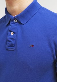 Detailní záběr osoby v modrém polo tričku s límečkem, na kterém je na hrudi malé logo s červenými, bílými a modrými detaily.