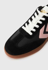 Svart sneakers med en kombinasjon av semsket skinn og mesh-materialer, hvite snørebånd, rosa sikksakk-detaljer og en brun gummisåle.