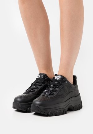 Pieds chaussés de baskets compensées noires à semelle épaisse avec un motif floral en relief et le logo Guess sur la languette ainsi qu'un détail métallique à l'avant.