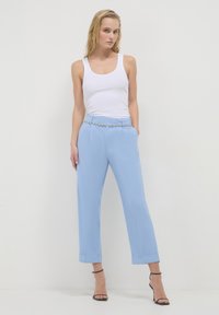 Donna in piedi contro uno sfondo bianco, indossa una canotta bianca, pantaloni azzurri chiari con una cintura decorativa e sandali neri con tacco alto.