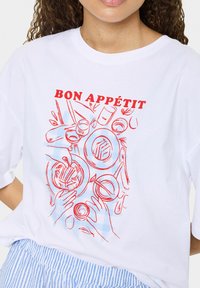 Valkoinen t-paita, jossa on punainen käsin piirretty design ruoka-esineistä ja käsistä, ja jossa lukee "BON APPÉTIT" selkeästi yläpuolella.