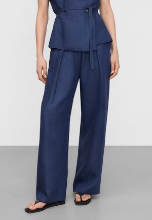 Persoon die marineblauwe wijde broek en bijpassende top met riem draagt, staat op blote voeten op een grijze vloer tegen een witte achtergrond.