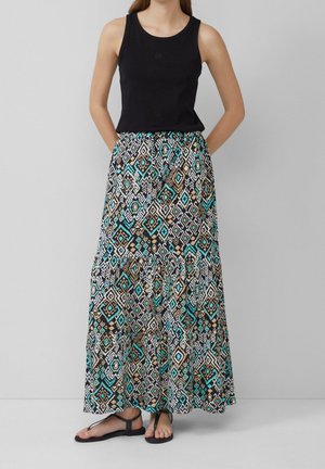 Kvinde iført sort ærmeløs top, lang nederdel med geometrisk mønster i turkis, sort, hvid og beige, og sorte sandaler, stående mod grå baggrund.