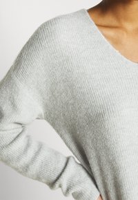 Světlé šedé pletené svetr s výstřihem do V, s měkkou strukturou a žebrovaným vzorem. Volný střih s dlouhými, mírně zúženými rukávy.