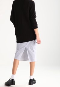 Osoba oblečená v černém oversized svetru, světle modré a bílé pruhované midi sukni s rozparkem na zadní straně, bílých ponožkách a černých společenských botách, stojí.