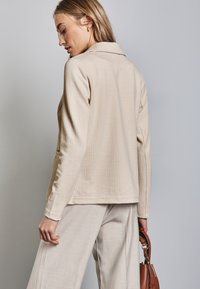 Beige teksturoidu bleiseri, jossa on lovettu kaulus ja pitkät hihat, sivutaskut ja rento leikkaus, yhdistettynä vaalean beigeihin housuihin.