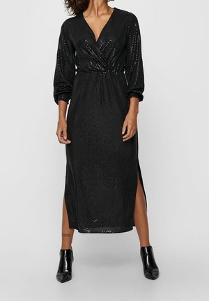 Kobieta ubrana w czarną, długorękawową sukienkę midi z cekinami i rozcięciami po bokach, w połączeniu z czarnymi botkami, stojąca na jednolitym tle.