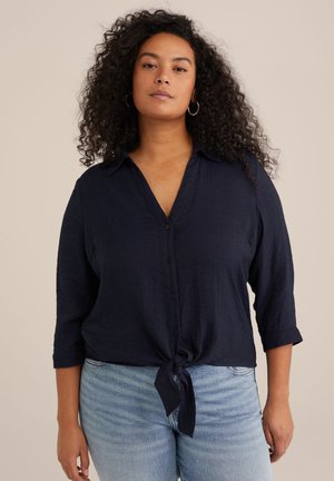 Camicia blu navy con bottoni, dettagli con nodo anteriore e tessuto testurizzato, dotata di colletto e maniche a tre quarti, abbinata a jeans chiari.