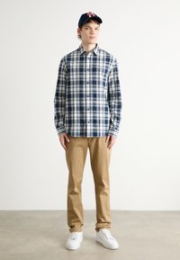 Camisa a cuadros en azul marino y blanco con diseño de botones, combinada con pantalones beige y zapatillas blancas, sobre un fondo liso.
