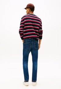 Debel mornarsko moder pulover z rdečimi in belimi črtami, kombiniran z modrimi kavbojkami in belimi supergami. Pogled od zadaj poudarja teksturo pletenega puloverja.