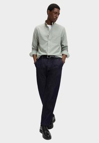 Camicia button-up verde chiaro con taschino sul petto, abbinata a pantaloni in denim scuri e scarpe nere, creando un look casual ma allo stesso tempo strutturato.