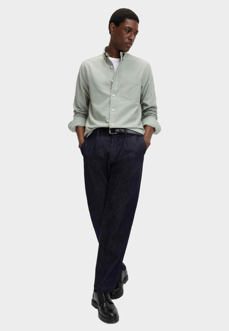 Camicia button-up verde chiaro con taschino sul petto, abbinata a pantaloni in denim scuri e scarpe nere, creando un look casual ma allo stesso tempo strutturato.