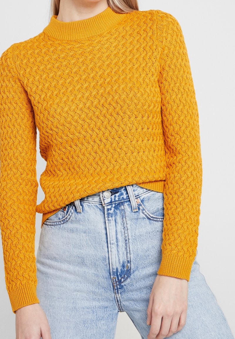 Femme portant un pull en maille texturée jaune moutarde associé à un jean en denim bleu clair taille haute.