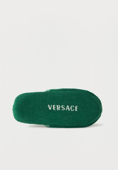 Πράσινο παντόφλα από πετσέτα με τη λέξη "VERSACE" κεντημένη με λευκά γράμματα στη σόλα, εμφανιζόμενη από κάτω σε λευκό φόντο.