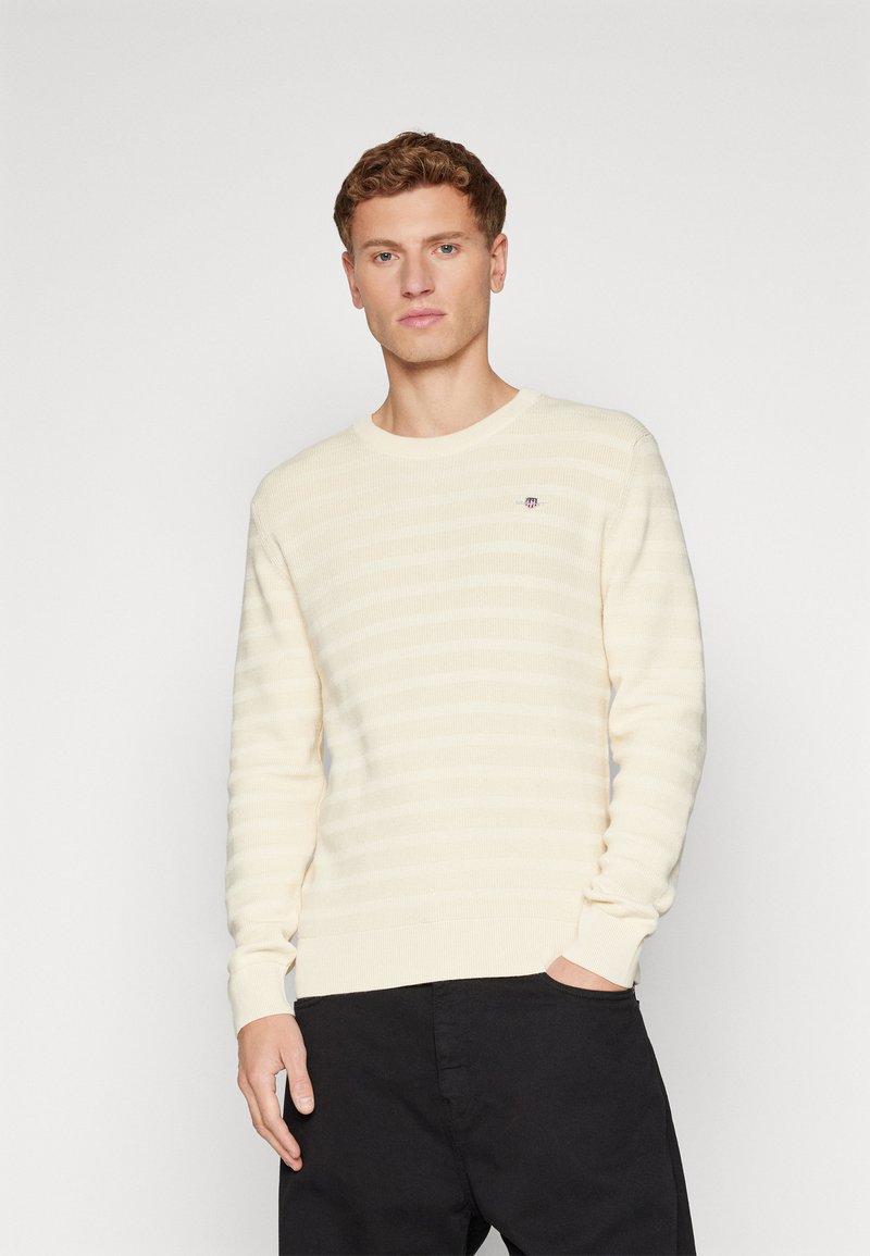 Młody mężczyzna z kręconymi włosami, ubrany w sweter w kremowe paski i czarne spodnie, stoi z jedną ręką w kieszeni na jednolitym tle.