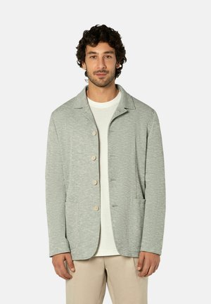 Man die een lichtgrijs jasje met knopen draagt over een wit overhemd en beige broek, staand tegen een effen witte achtergrond.