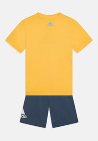 Keltainen puuvillapaita lyhyillä hihoilla ja pyöreällä kaula-aukolla, yhdistettynä tummansinisiin shortseihin, joissa on valkoinen Adidas-logo ja geometrinen koriste.