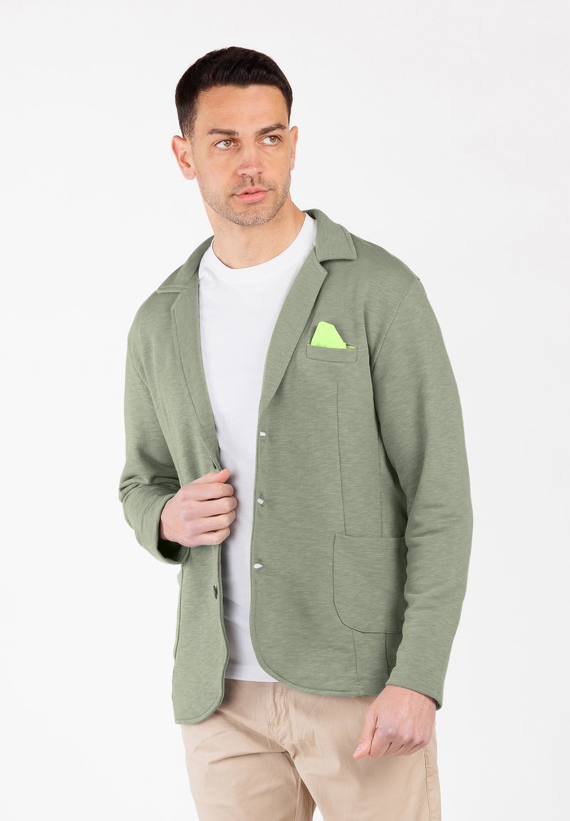Hombre con blazer casual verde claro, pañuelo de bolsillo neón, camiseta blanca y pantalones beige, mirando ligeramente hacia un lado.