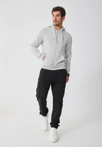 Felpa con cappuccio grigia chiaro con cerniera e tasche a marsupio, abbinata a pantaloni cargo neri con tasche laterali e sneakers bianche.