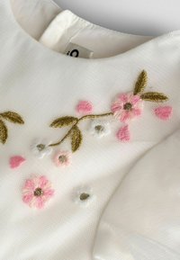 Weißer Stoff mit strukturierter Oberfläche und bunter Blumenstickerei in Pink, Weiß und Gold. Rundhalsausschnitt ist sichtbar.